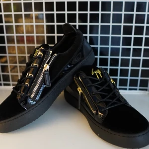 Svarta sneakers från Giuseppe Zanotti - Snygga svarta sneakers från Giuseppe Zanotti med lyxiga gulddetaljer, dragkedjor på sidorna och signaturlogo på plösen. Skorna har en mix av mocka och skinn, platt sula och snörning. Perfekta för dig som vill sticka ut med exklusiv streetstyle. Str 41, pris går att diskutera 