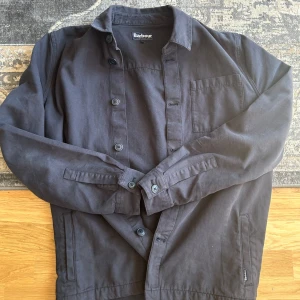 Mörkblå overshirt från Barbour - Sjukt stilig overshirt från Barbour med klassisk krage, raka linjer och knäppning framtill. Jackan har en bröstficka och två sidofickor. Perfekt för dig som vill ha en uppklädd men ändå soft stil. Passar lika bra utomhus som inomhus! Plagget är knappt använt och har riktigt fint material. Nypris på cirka 2300kr! 