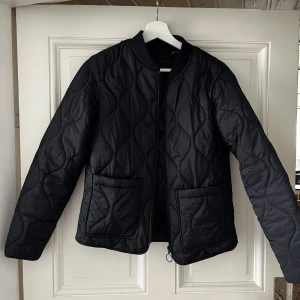 Svart Jacka - Svart jacka med rundad krage och två stora fickor framtill. Jackan har en snygg dragkedja med ring och vågformat quiltmönster över hela ytan. Perfekt för dig som vill ha en stilren och trendig jacka till vardags.