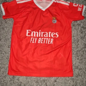 Röd Benfica fotbollströja Adidas - Snygg röd Benfica fotbollströja från Adidas med vita detaljer och klubbmärke på bröstet. Tröjan har korta ärmar, V-ringad krage och tryck från Emirates och Sagres. Tillverkad i lätt och ventilerande polyester, perfekt för match eller träning.