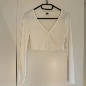Vit croppad topp med knappar från SHEIN - Säljer en vit croppad topp från SHEIN i ribbat material. Toppen har lång ärm, v-ringning och knappar framtill. Perfekt att styla med höga jeans eller kjol för en clean look. Aldrig använd