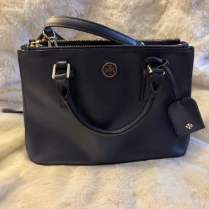 Fin marinblå handväska från Tory Burch - Stilren mörkt marinblå handväska från Tory Burch i skinn med guldfärgade metalldetaljer och dubbla handtag. Väskan har dragkedja upptill, avtagbar axelrem och logga framtill. Insidan är fodrad med mönstrat tyg och har flera fack för smart organisering.