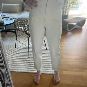 Vita högmidjade kostymbyxor - Snygga vita kostymbyxor med hög midja och croppad längd. Byxorna har raka ben, pressveck och diskreta fickor framtill. Perfekta för en clean och stilren look. Materialet känns mjukt och har en lätt stretch för extra komfort.