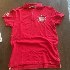Röd polo t-shirt - Tommy Hilfiger  - Röd polo t-shirt från Tommy Hilfiger med coolt USA-inspirerat tryck och stjärnor på bröstet. Klassisk krage med två knappar och detaljer på ärmen. 