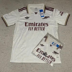 Vit Arsenal fotbollströja Adidas M - Snygg vit Arsenal fotbollströja från Adidas med vinröda detaljer och klubbens logga på bröstet. Tröjan har korta ärmar, klassisk krage med ränder och tryckta sponsordetaljer. Tillverkad i lätt och ventilerande material, perfekt för match eller träning.