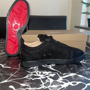 Christian Louboutin svarta sneakers mocka - Exklusiva svarta sneakers från Christian Louboutin i mjuk mocka med klassisk röd sula och broderad logga baktill. Skorna har rund tå, snörning och diskreta metalldetaljer på sidan. Insidan är i beige skinn för extra komfort. Perfekt för dig som gillar lyxiga detaljer.