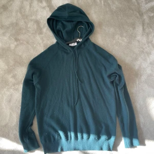 Mörkgrön hoodie i kashmir från Tiger of Sweden - Mysig mörkgrön hoodie från Tiger of Sweden i lyxig kashmir. Tröjan har huva med snörning och långa ärmar. Perfekt för dig som gillar en stilren och avslappnad look med premiumkänsla.