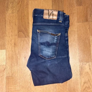 Mörkblå jeans från Nudie Jeans, 27/32 - Säljer ett par mörkblå jeans från Nudie Jeans i klassisk femficksmodell. Jeansen har kontrastsömmar, mellanblå tvätt och Nudie-logga på bakfickan. Modellen är slim och materialet är mjukt denim i bomull. Perfekta för dig som gillar stilrena och tidlösa jeans.