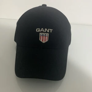 Svart keps från GANT med broderad logo - Svart keps från GANT med klassisk böjd skärm och broderad GANT-logo samt sköld med stjärnor och ränder framtill. Snygg och stilren design som passar till det mesta. Justerbar baktill för perfekt passform.