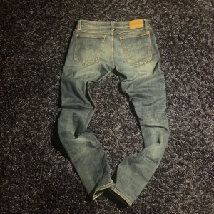 Nudie Jeans blå jeans W30 L34 - Snygga blå jeans från Nudie Jeans med klassisk femficksdesign och orangea kontrastsömmar. Jeansen har en rak passform och subtila slitningar för en cool look. Tillverkade i bomull med broderad logga på bakfickan och läderpatch i midjan. Modell , tight long John