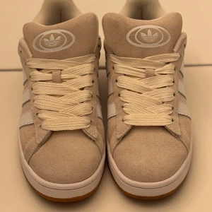 Beige Adidas Campus sneakers mocka - Snygga Adidas Campus sneakers i beige mocka med klassiska vita ränder på sidorna och vit logga på plösen och hälen. Skorna har platt sula i gummi, rund tå och breda vita snören. Perfekt för dig som gillar stilrena och tidlösa sneakers med retrovibe.