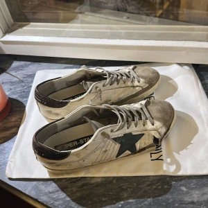 Golden Goose Superstar sneakers grön/vit - Golden Goose Superstar sneakers i vitt skinn med grön stjärna på sidan och detaljer i mocka. Hälpartiet har ett mönstrat parti i mörkare färg. Klassisk låg modell med snörning och platt sula. Snyggt slitna för en cool streetstyle-look. Men högra skon har ett hål på sidan