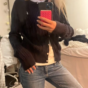 Mörkblå kabelstickad kofta Polo Ralph Lauren - Kabelstickad kofta från Polo Ralph Lauren med klassisk passform. Koftan har vita knappar framtill, ribbade muddar och den ikoniska broderade loggan på bröstet. Perfekt att slänga över en t-shirt eller skjorta för en clean look. Står att storleken är XL men antar att det är för barn då 