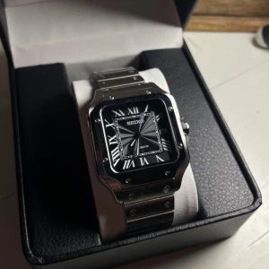 Seiko Santos mod - Helt nybyggd Seiko Santos Mod  Specifikationer: Storlek: 38mm Urverk: Nh35 (Automatiskt)  Material: 904L Rostfritt Stål Glas: Sapphire Glas Datumvisare Boett: Fyrkantig Tavla: Svart/grå Armband: Butterfly lås Krona: blå kristall Sten  Vattentålighet: 30 Meter Klocklåda ingår