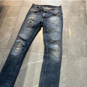 Nudie Jeans blå slitna jeans 29/32 - Säljer ett par blå jeans från Nudie Jeans med snygga slitningar och lagom tvättad look. Klassisk femficksmodell med orange sömmar och broderad bakficka. Jeansen är raka i modellen och har normal passform. Perfekta för dig som gillar en avslappnad stil.