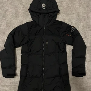 Svart lång dunjacka Sail Racing - Svart lång dunjacka från Sail Racing med huva och dragkedja framtill. Jackan har logga på bröstet och ärmen, samt justerbara muddar med kardborre. Perfekt för kalla dagar när du vill hålla värmen och ändå se stilren ut. Använd i en vinter.