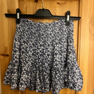 Blommig blå volangkjol - Supersöt kort kjol i mörkblått med vitt blommönster och volangkant. Kjolen är även väldigt stretchig 