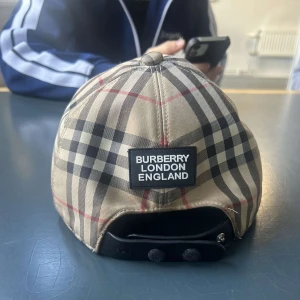 Burberry rutig keps med klassiskt mönster - Säljer en ikonisk keps från Burberry med det klassiska rutiga mönstret i beige, svart och rött. Kepsen har justerbar rem bak och en tydlig Burberry London England-patch. Tillverkad i bomull och polyester för en skön passform.