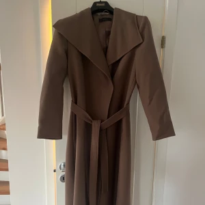 Brun kappa från Ellos med bälte - Snygg brun kappa från Ellos med bred krage och knytbälte i midjan. Lång modell med raka ärmar och stilren design. Lite tunnare i materialet så passar perfekt nu till våren och hösten. Knappt använd och har inga defekter. Nypris 1000kr