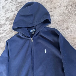 Marinblå ralph Lauren zip - Snygg mörkblå hoodie från Polo Ralph Lauren med dragkedja och klassisk vit logga på bröstet. Tröjan har huva, långa ärmar och en stor ficka framtill. Perfekt för en chill och stilren look.