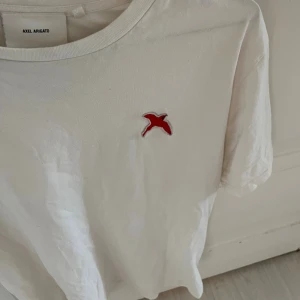 Vit t-shirt från Axel Arigato - Vit t-shirt från Axel Arigato med röd broderad fågel på bröstet. Klassisk rund halsringning och korta ärmar. Skön och stilren modell i mjuk bomull, perfekt till jeans eller shorts.