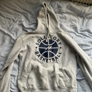 SoleStory Basketball Hoodie - Grå hoodie från SoleStory med stort blått baskettryck och texten 'SoleStory Basketball' på framsidan. Huva med snörning och känguruficka. Liten blå basketkorg med S på ryggen. Perfekt för dig som gillar basket och streetwear. Storlek M passar även S
