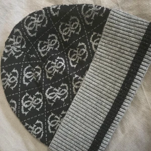 Svart och grå stickad mössa från Guess - Snygg stickad mössa från Guess i svart och grått med logomönster över hela mössan. Uppvikt kant i ribbstickat grått med en svart rand och diskret glittereffekt. Perfekt accessoar för att hålla värmen och samtidigt se trendig ut.