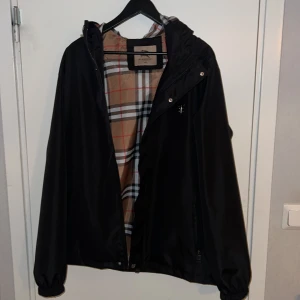 Svart vindjacka från Burberry XXL (passar mer som L) - Svart vindjacka från Burberry i storlek XXL med klassiskt rutigt foder i beige, rött, vitt och svart. Jackan har huva, dragkedja framtill och elastiska muddar. Perfekt för dig som vill ha en stilren jacka med ikoniska Burberry-detaljer. Storlek är XXL men den passar bättre folk som är XL eller L 