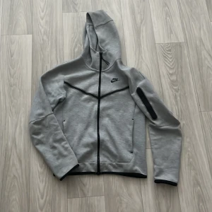 Grå Nike hoodie med dragkedja XS - Nike Tech Fleece hoodie i ljusgrått med svarta detaljer och hel dragkedja framtill. Tröjan har huva, lång ärm med ficka och dragkedja på ena ärmen samt två sidofickor. Perfekt för chill eller träning.