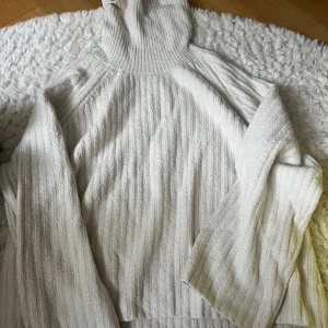 Ljus polotröja i ribbad stickad bomull - Mysig ljus polotröja med ribbad struktur och lång ärm. Tröjan är stickad i mjuk bomull och har en hög, bred polokrage som ger en avslappnad och trendig look. Perfekt för lager-på-lager under kyliga dagar.