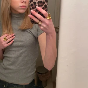 Grå ribbad polotopp - Säljer en grå ribbad polotopp med korta ärmar och tight passform. Toppen har en klassisk polokrage och är tillverkad i ett mjukt, stretchigt material som sitter snyggt på kroppen. Perfekt att styla med jeans eller kjol för en clean look.