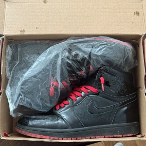 Nike Air Jordan 1 Svart/Röd Highs - Säljer ett par Nike Air Jordan 1 highs i svart med röda detaljer och röda skosnören finns även svarta som ingår med i kartongen. Skorna har en klassisk siluett med hög modell och är tillverkade i skinn med glansiga partier. Perfekta för dig som gillar streetwear och sneakers med attityd.använda tre-fyra gånger 