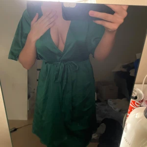 Grön omlottklänning med knytband - Säljer en snygg grön omlottklänning med djup v-ringning och knytband i midjan. Klänningen har korta ärmar och är tillverkad i ett glansigt, mjukt material som ger en lyxig känsla. Perfekt för dig som vill ha en chic och trendig look.