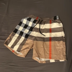 Burberry rutiga badshorts med snörning - Snygga Burberry badshorts med klassiskt rutigt mönster i beige, svart, vit och röd. Shortsen har elastisk midja med snörning och meshfoder på insidan. Perfekt för sommaren och strandhäng, med en bakficka och cool design.