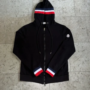 Svart Moncler hoodie med logga - Svart hoodie från Moncler med hel dragkedja framtill och Moncler-logga på ärmen, Ränder i rött, vitt och blått på huvan och ärmsluten. Storlek S och nytt skick, fraktar inom 24h✅