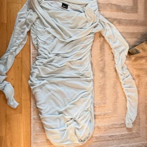 Ljusblå omlottklänning från Gina Tricot - Säljer en ljusblå kortklänning från Gina Tricot med snygg omlottkrage och rynkade detaljer längs hela klänningen. Materialet är mjuk mesh med ett tunt foder under. Klänningen har långa ärmar och sitter snyggt figurnära. Perfekt för dig som gillar trendiga och stilrena plagg.