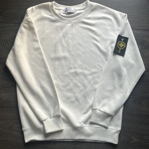 Vit sweatshirt från Stone Island - En knappen är av,Vit sweatshirt från Stone Island med klassisk rund hals och ribbade muddar. Tröjan har den ikoniska Stone Island-loggan på vänster ärm. Tillverkad i mjuk bomull för en skön och avslappnad stil. Perfekt för dig som gillar stilrena och trendiga plagg.