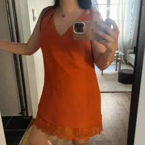Orange klänning från Sandro  - Superfin orange kort klänning från Sandro med v-ringning fram och snygg öppen rygg med banddetalj. Klänningen har en volangkant nertill och är ärmlös. Materialet är polyester och känns lätt och bekvämt. Perfekt för dig som gillar färg och vill sticka ut. Oanvänd!!