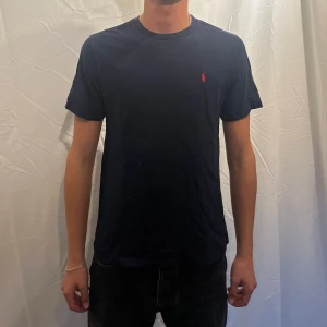  t-shirt från Ralph Lauren - Snygg mörkblå t-shirt från  Ralph Lauren med klassisk röd logga broderad på bröstet. T-shirten har rund halsringning och korta ärmar. Tillverkad i mjuk bomull för en skön känsla och clean look. Storleken är L i barn men motsvara S i herr. Modellen 180cm