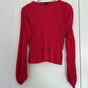 Röd smockad blus med volang - Supersöt röd blus från Gina Tricot med smockad midja och volangkant nedtill. Långa puffiga ärmar med resår vid handlederna och rund halsringning. Tunt, strukturerat tyg som ger en snygg och trendig look.