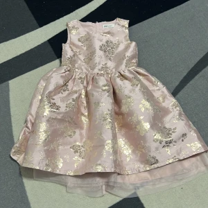 Rosa blommig klänning från H&M - Supersöt rosa klänning från H&M med glansigt tyg och guldiga blommor. Klänningen är ärmlös, har rund halsringning och ett fluffigt lager av tyll under kjolen för extra volym. Dragkedja bak för enkel påklädning. För barn 