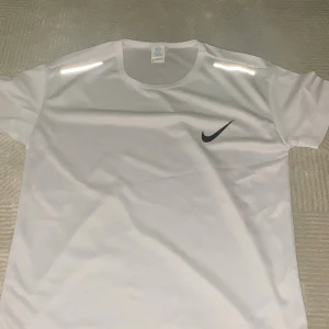 Vit Nike t-shirt med svart logga Reflection - Fräsch vit t-shirt från Nike med klassisk svart swoosh på bröstet. T-shirten har rund hals, korta ärmar och är gjord i ett lätt polyestermaterial som andas. Perfekt för träning eller chill. Snygga reflekterande detaljer på axlarna.de är helt oanvända!