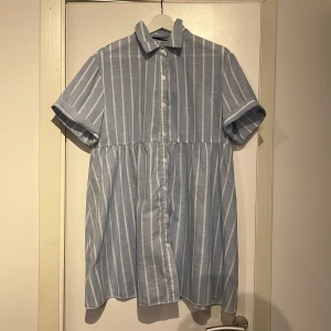 Blåvit randig skjortklänning - Superfin blå och vit randig skjortklänning med korta ärmar och klassisk krage. Klänningen har knappar hela vägen framtill och är tillverkad i ett lätt och luftigt tyg, perfekt för varma dagar. Avslappnad passform och snyggt fall.