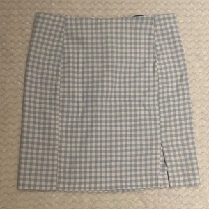 Blåvit rutig kortkjol med slits - Supersöt kortkjol i blåvitt rutigt mönster med liten slits framtill. Kjolen har en rak passform och dragkedja bak. Perfekt för dig som gillar en preppy och fräsch stil. Materialet känns som bomull eller polyester, lätt och skönt för varmare dagar.