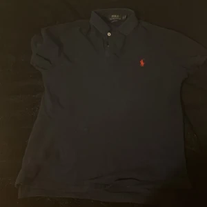 Mörkblå Polo Ralph Lauren t-shirt - Snygg mörkblå t-shirt från Polo Ralph Lauren med klassisk krage och två knappar framtill. Den har en röd broderad logga på bröstet och är i en slim fit-modell. Perfekt för dig som gillar stilrena och tidlösa plagg.