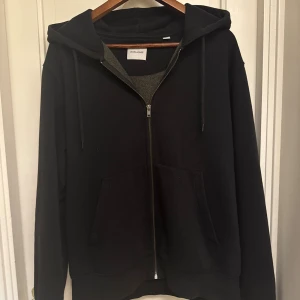 Svart hoodie från Jack & Jones M - Svart hoodie från Jack & Jones i storlek M. Klassisk modell med dragkedja framtill, huva med snörning och två stora fickor. Mjuk insida och normal passform, perfekt för chill dagar eller när du vill ha en enkel stil.