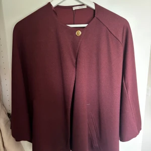 Vinröd kappa från Zara med guldknapp - Stilren vinröd kappa från Zara med en snygg guldknapp vid halsen. Jackan har en rak och lös passform med trekvartsärmar och diskreta fickor framtill. Perfekt för dig som gillar minimalistisk och elegant stil.