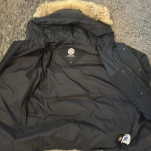 Svart Canada Goose dunjacka med päls BRA PRIS! - Säljer en svart äkta Canada Goose long parka med avtagbar huva och beige pälsdetalj. Jackan har klassisk patch på ärmen, flera fickor och dragkedja med knappar. Perfekt för kalla vinterdagar och riktigt varm. Snygg och tidlös modell. Det finns inte bättre på marknaden än detta för samma pris!