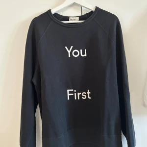 Svart Acne Studios Tröja - Svart sweatshirt från Acne Studios med vit text 'You First' på framsidan. Storlek L passar även M. Tröjan har en klassisk rund halsringning och långa ärmar. Tillverkad i mjuk bomull för en skön och avslappnad look.