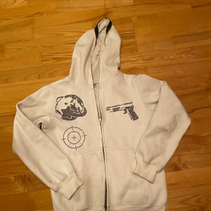 Vit hoodie Aim For The Moon  - Vit hoodie med dragkedja från Aim For The Moon. Framsidan har tryck med pistol, sikte och måne. Utrustad med huva och ribbade muddar. Perfekt för dig som gillar streetwear och vill sticka ut med coola detaljer. Fullzip och är limited edition 